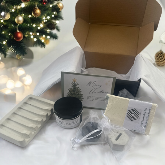 Christmas gift set with candles, cards, and other items on a white surface with a decorated tree in the background.